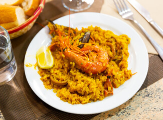 Hearty and healthy lunch - paella with seafood, decorated with large shrimp. Main course is served with pieces of baguette and glass with drink
