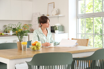 Young woman with cup of coffee reading magazine at table in dining room