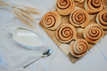 Fresh cinnabons on a wooden tray along with milk and spoon on a white fabric background