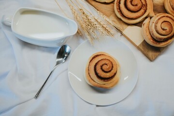 Fresh cinnabon on a plate on a background of other buns on a wooden tray and milk with a spoon 