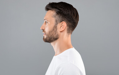 Profile portrait of middle-aged bearded man in white t-shirt over grey studio background, copy space. Side view of handsome confident man posing on gray, standing straight and looking aside