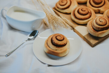 Fresh cinnabon on a plate on a background of other buns on a wooden tray and milk with a spoon