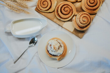 Fresh cinnabon on a plate on a background of other buns on a wooden tray and milk with a spoon 