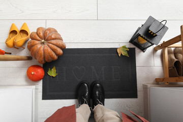 Woman with autumn decor and lantern standing near doormat on white wooden floor, top view