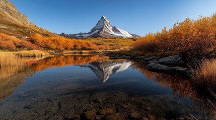 Fototapeta premium Matterhorn mountain rises sharply with a snow-covered peak, mirrored in a tranquil lake, embraced by fiery autumn colors and a clear blue sky above