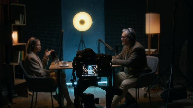 Medium full arc shot of digital camera on tripod recording podcast in studio, with Caucasian man and woman debating on business environment and challenges, on teal blue background