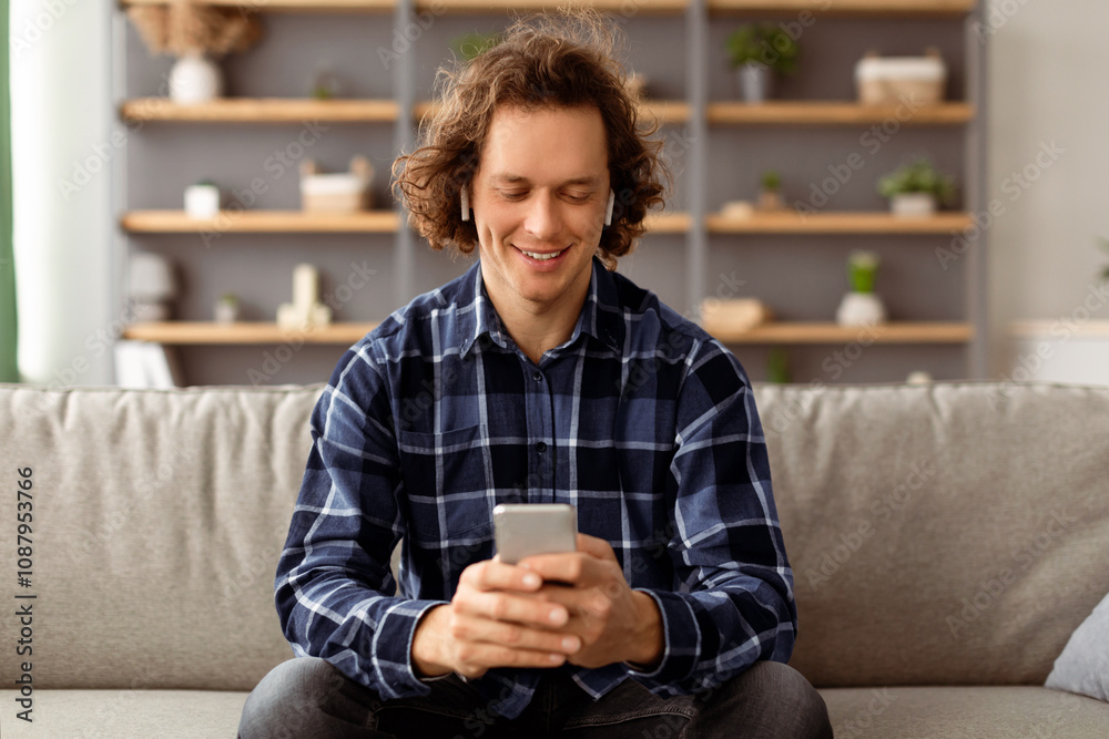 Poster Positive Guy In Earbuds Using Smartphone Listening To Music Online Sitting On Sofa At Home. Man Relaxing Using Mobile Application And Browsing Internet In Living Room Indoor.