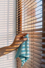 Cleaning wooden blinds with a blue cloth in bright sunlight, creating shadows indoors