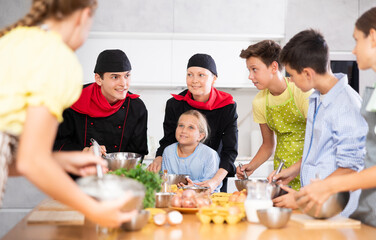 Young guy and adult woman cook in uniform teaches group of children how to cook dish
