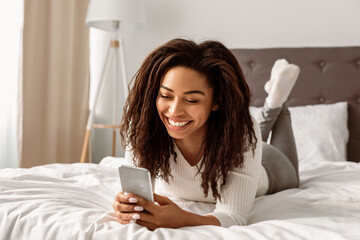 Mobile Phone User. Portrait of happy African American woman lying in bed and smiling while reading text message from someone, holding and using smartphone, chatting with lover or browsing social media