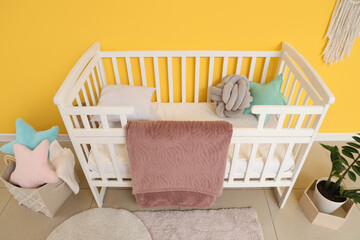 Interior of stylish children's bedroom with crib, cozy blanket and houseplant near yellow wall