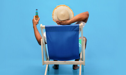 Back view of young black guy sitting in lounge chair with bottle of beer, enjoying summer vacation,...