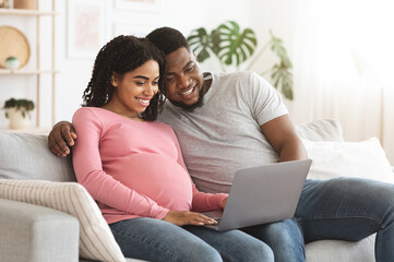 Pregnant african couple having fun at home, using laptop together, watching funny videos, empty space