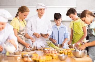 Enthusiastic young chef giving culinary classes to group of interested curious tween girls and boys, teaching to cook deliciously..