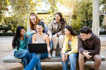 Medium shot of a group of 6 young people between 20 and 30 years old of different ethnicities sitting on a park bench using a laptop.Concept of online shopping, working from the street.