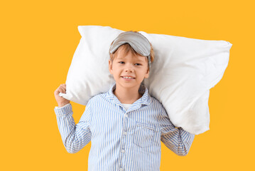 Cute little happy boy in pajamas with sleeping mask and pillow on yellow background