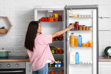 Young Asian woman taking jar of preserves from open fridge full of fresh food in kitchen