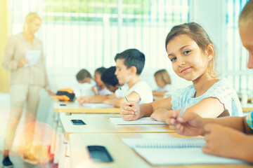 Portrait of positive small female pupil sitting at desk studying in classroom