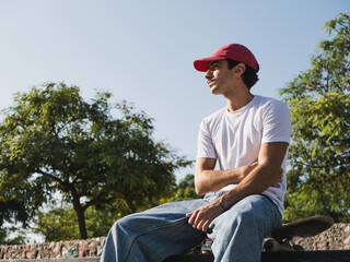 Young man in red cap relaxing on skateboard in urban park