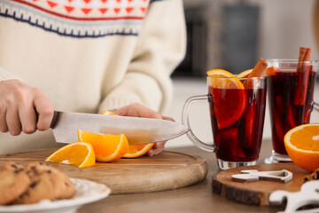Young woman in warm sweater with glass cups of hot mulled wine cutting orange in kitchen, closeup