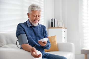 Senior man measuring blood pressure on sofa indoors
