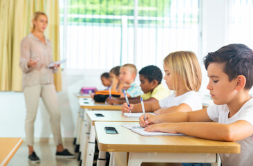 Diligent tweens studying in classroom, listening to female teacher and writing in notebooks