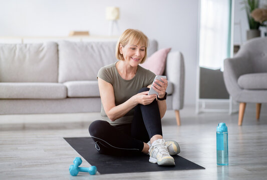 Fit senior woman relaxing on yoga mat with smartphone after her home workout, checking new sports videos online. Mature Caucasian lady taking break after yoga practice. Active lifestyle concept