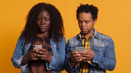 African american sweet life partners laugh at funny videos on mobile phone apps in studio, enjoying leisure time together against yellow background. Cheerful romantic couple use smartphone. Camera A.