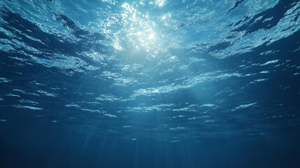 A large body of water under a cloudy blue sky with sun shining through the clouds over the water and the ocean below, with a bright blue sky and white clouds.