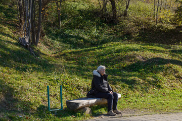 Elderly woman is sitting on log bench in Skhidnytsia, Ukraine.