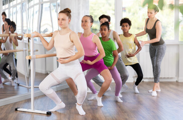 Obraz premium Motivated teenage girl learning grand plie at barre during group ballet class for beginners under guidance of experienced female instructor in sunlit studio