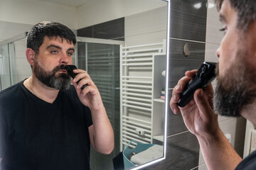 Man shaving his beard in the bathroom. Beard care, electric razor.