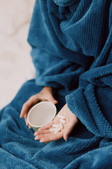Person wrapped in blue blanket holding cup of water and pill in one hand, another cup of water in the other