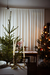 Long beige white candles in a candlestick with brown velvet bows. Christmas trees and fir tree branches on a brown wooden table