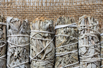 A top view image of several white sage smudge sticks lined up on a ripped burlap sack cloth.