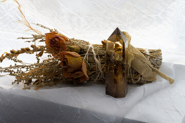 A close up image of a small smoky citrine crystal tower on a white window ledge with a decorative rose and sage smudge stick.