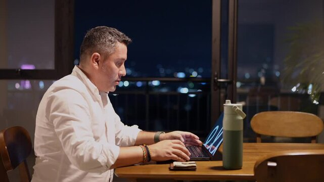 Hombre trabajando en mesa de comedor al lado del balc&oacute;n en la noche