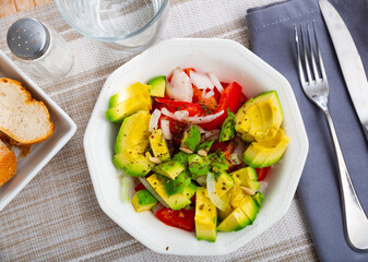 Avocado, tomato, onion salad served on table in blue plate with serving pieces.