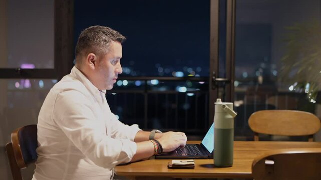 Hombre trabajando en mesa de comedor al lado del balc&oacute;n en la noche