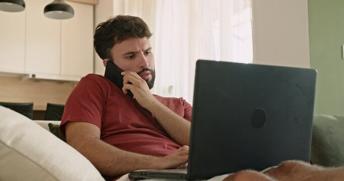 A young Caucasian man sits on his couch at home, working on his laptop and talking on his phone. He appears to be multitasking and handling business or personal matters.