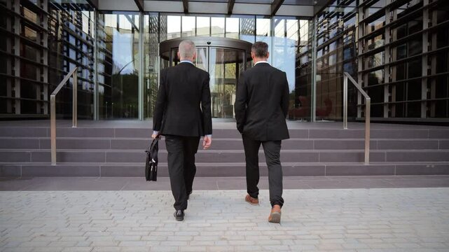 Rear view of two distinguished business executives walking confidently together towards modern corporate building entrance after successful meeting, showcasing professional partnership