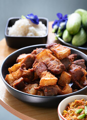 Deep fried sliced crispy pork belly in a black plate with chili sauce and sticky rice, assorted vegetables in background such cucumber and butterfly pea flower.