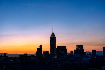 The Mexico City skyline at sunset, featuring Torre Latinoamericana and other buildings silhouetted against a colorful orange and blue sky.