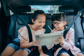 A pair of Asian sisters sit side by side in a car and use digital tablets. They looked happy, engaged, and enjoying their devices on the go.