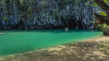 A sheer cliff above a calm turquoise river. Canoes with tourists sail towards the dark entrance of...