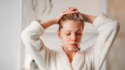 Beauty practice. Treatment procedure. Attractive young woman wearing white soft robe tying hair with hands with barrette in mouth.