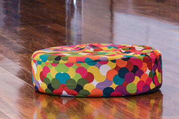 A vibrant multicolored pouf on a polished hardwood floor, with a softly blurred background. The bright colors contrast elegantly with the warm wood tones, creating a cozy ambiance.