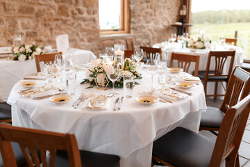 Festive round table is ready for the guests. Wedding table for guests with white tablecloth, plates, glasses, souvenirs, decorated with flowers and chairs around. Elegant dining table without people