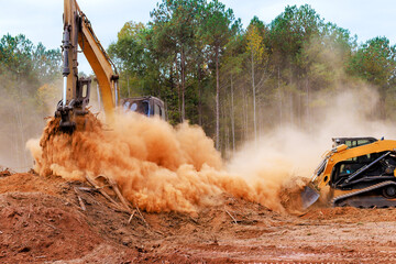 Heavy bulldozer machinery excavator works to clear land in wooded area, creating large clouds of...