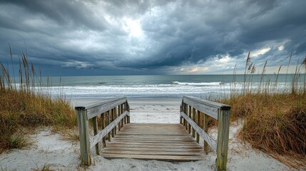 Wooden walkway leading to a serene beach and ocean, framed by tall grass and dramatic overcast skies, creating a peaceful coastal scene.